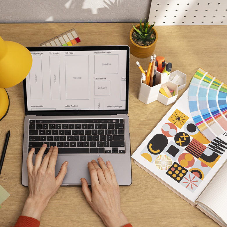 Diseñadora gráfica trabajando en formatos de piezas publicitarias en su notebook, junto a un muestrario de colores impreso